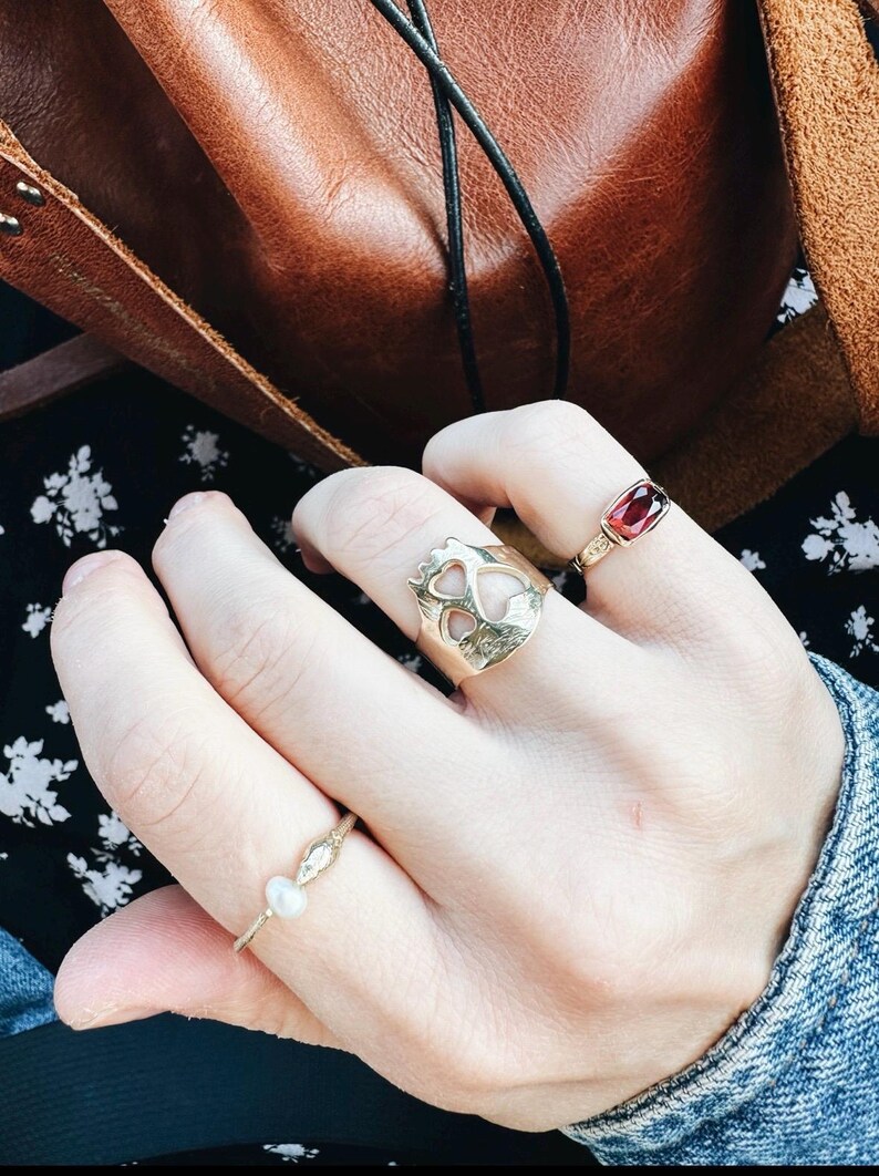 Op de afbeelding: Een hand met drie gouden ringen. De ring op de wijsvinger heeft een uitgesneden ontwerp van twee harten. De ring op de middelvinger heeft een rode edelsteen. De ring op de pink heeft een kleine parel.