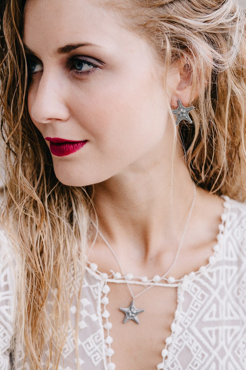 May include: A woman wearing a white lace top with a silver necklace and earrings featuring a starfish design.