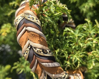 May include: A braided headband featuring a multi-textured design with strips of brown suede, black fabric, beige cord, and a gold-toned leopard print ribbon. The braided construction creates a thick, layered appearance with alternating colors and materials.