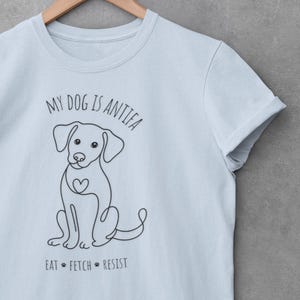 Puede incluir: Camiseta azul claro con el texto "MY DOG IS ANTIFA" sobre un dibujo lineal de un perro con un corazón en el pecho. Debajo del perro está el texto "EAT * FETCH * RESIST". La camiseta tiene mangas cortas y está colgada de una percha de madera.