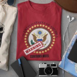 May include: Red t-shirt featuring the United States of America seal with a "WARNING CONTAINS NUTS" label. The shirt is surrounded by a tote bag, sunglasses, a notebook, a camera, a phone, a map, and a hat.