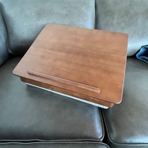 May include: A wooden laptop desk with a brown finish. The desk has a raised edge to prevent items from sliding off and a cushioned base. The desk is designed to be used on a lap or other surface.