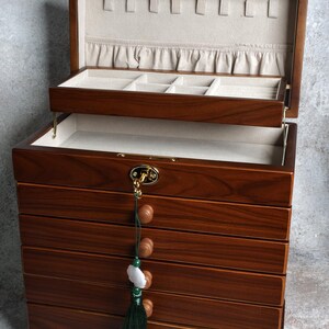 May include: A wooden jewelry box with multiple drawers and compartments. The box is made of dark brown wood and has a light-colored interior. A green tassel with wooden beads and a white stone hangs from the front. The box is open, revealing the interior.