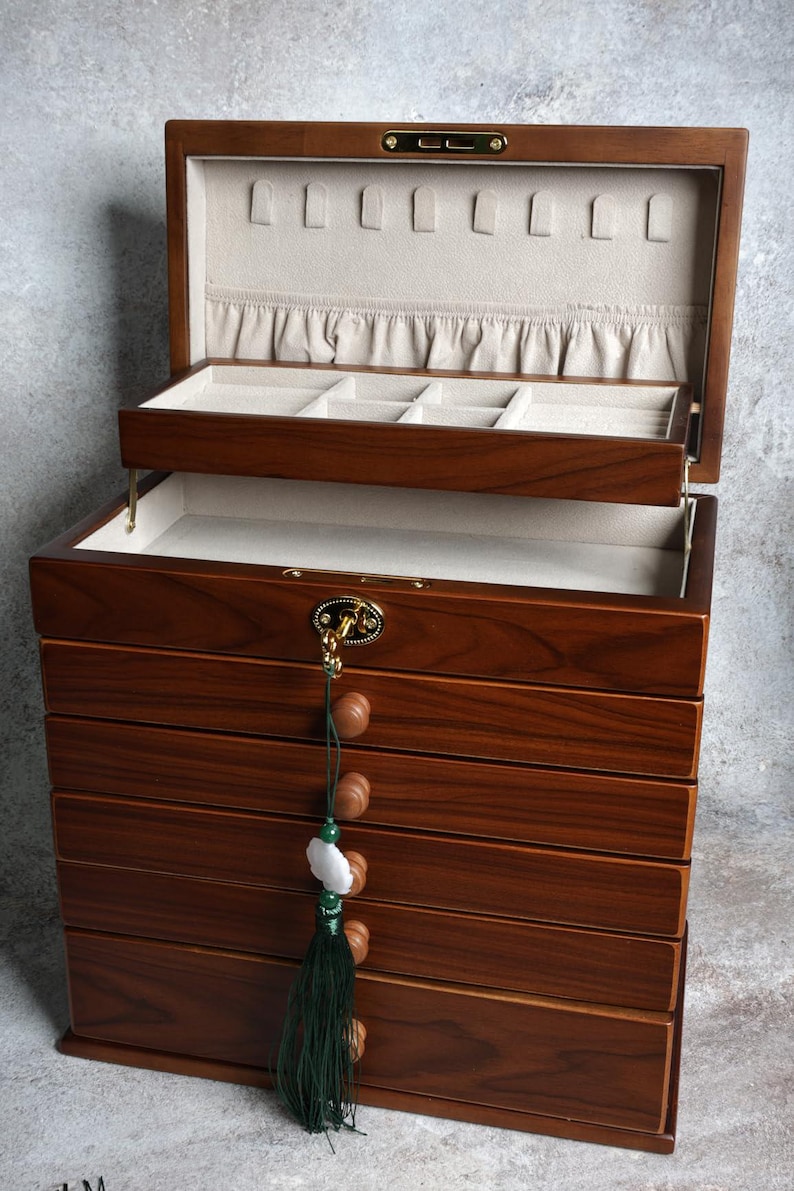 May include: A wooden jewelry box with multiple drawers and compartments. The box is made of dark brown wood and has a light-colored interior. A green tassel with wooden beads and a white stone hangs from the front. The box is open, revealing the interior.