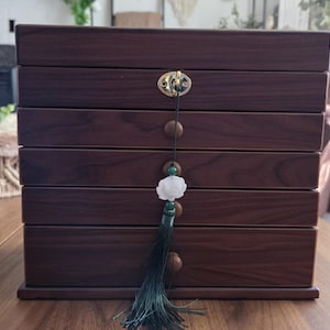 May include: A dark brown wooden jewelry box with six drawers. The box has a gold-colored clasp and a decorative tassel with a white flower pendant. The box is sitting on a wooden surface.
