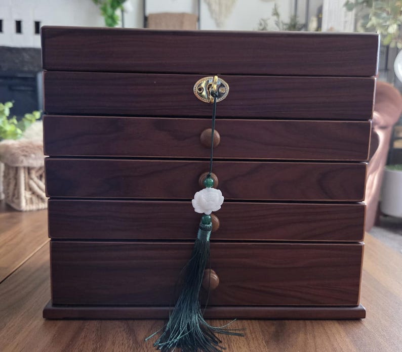 May include: A dark brown wooden jewelry box with six drawers. The box has a gold-colored clasp and a decorative tassel with a white flower pendant. The box is sitting on a wooden surface.