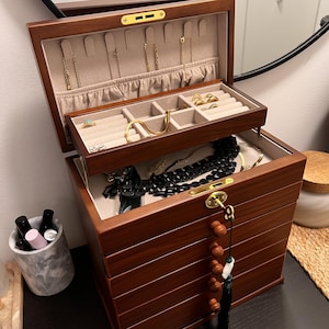 May include: A wooden jewelry box with multiple compartments and drawers. The box is open, revealing various necklaces, rings, and bracelets. The interior is lined with a beige fabric. The box has a key and a tassel.