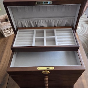 May include: A dark brown wooden jewelry box with multiple compartments and drawers. The interior is lined with a light gray fabric. The box is open, revealing ring slots and storage areas. A key is visible.