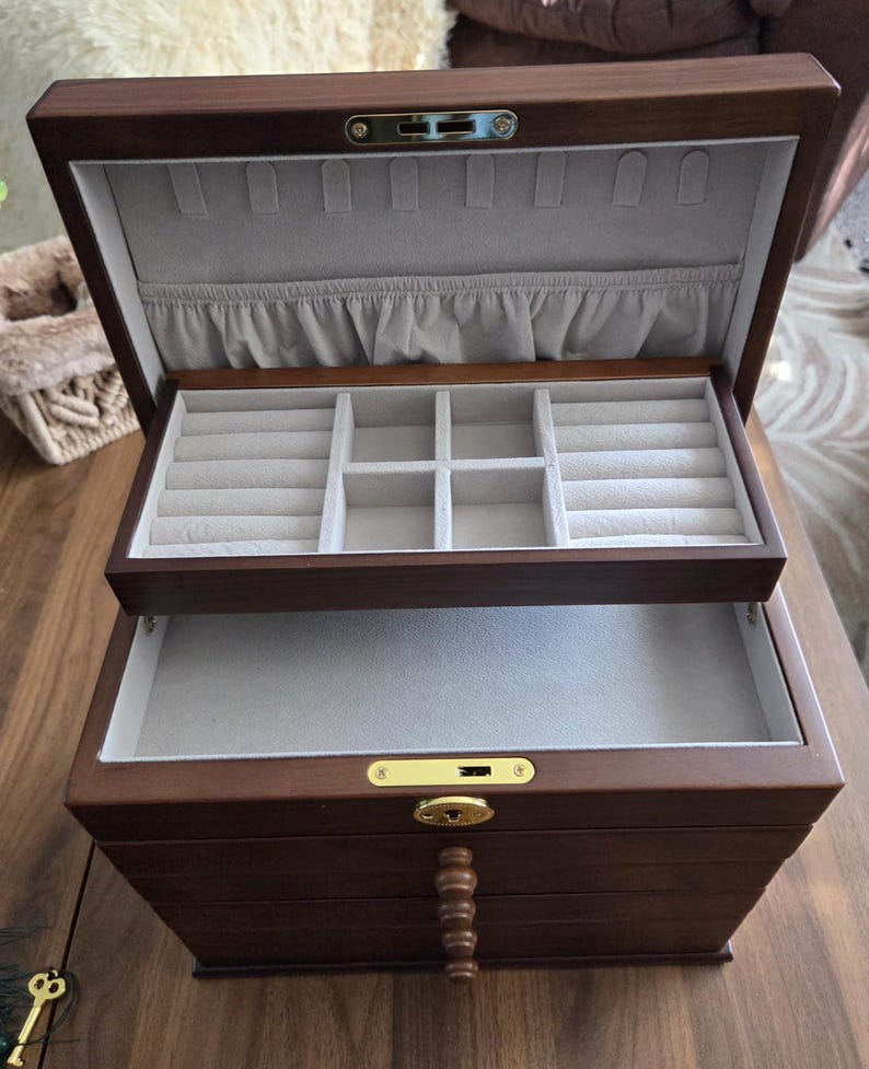 May include: A dark brown wooden jewelry box with multiple compartments and drawers. The interior is lined with a light gray fabric. The box is open, revealing ring slots and storage areas. A key is visible.