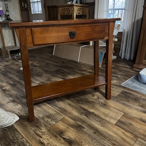 May include: A rectangular wooden console table with a medium brown finish, featuring a single horizontal drawer with a dark metal pull handle and a lower open shelf supported by four straight legs, showcasing a visible wood grain texture throughout.