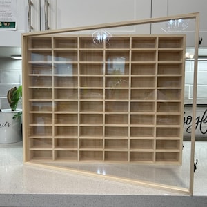 May include: A wooden display case with a clear glass door, featuring numerous small, evenly spaced compartments. The case is made of light-colored wood and is designed to showcase collectibles or small items. The door is partially open, revealing the interior.