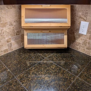 May include: Two wooden kitchen cabinets with ribbed glass doors are mounted under a dark brown cabinet. The cabinets are above a dark granite countertop. A white electrical outlet is visible on the right side of the image.