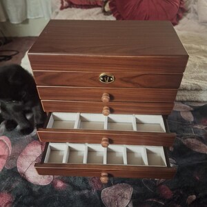 May include: A wooden jewelry box with multiple drawers, some of which are open, revealing compartments lined with a light-colored fabric. The box has a warm, reddish-brown finish and round wooden drawer pulls. A small, decorative metal emblem is on the top drawer.