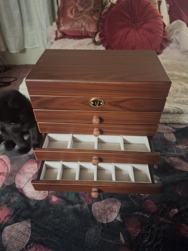 May include: A wooden jewelry box with multiple drawers, some of which are open, revealing compartments lined with a light-colored fabric. The box has a warm, reddish-brown finish and round wooden drawer pulls. A small, decorative metal emblem is on the top drawer.