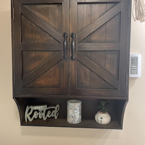 May include: A dark brown wooden cabinet with two doors and black handles. The doors have a diagonal cross design. Below the cabinet is a shelf with a decorative sign that says "Stay Rooted", a patterned vase, and a small potted plant.