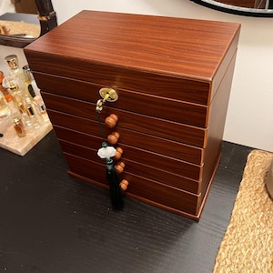 May include: A wooden jewelry box with a rich, reddish-brown finish. The box has multiple drawers and a decorative tassel with wooden beads and a jade-like ornament. A gold-colored clasp secures the lid.