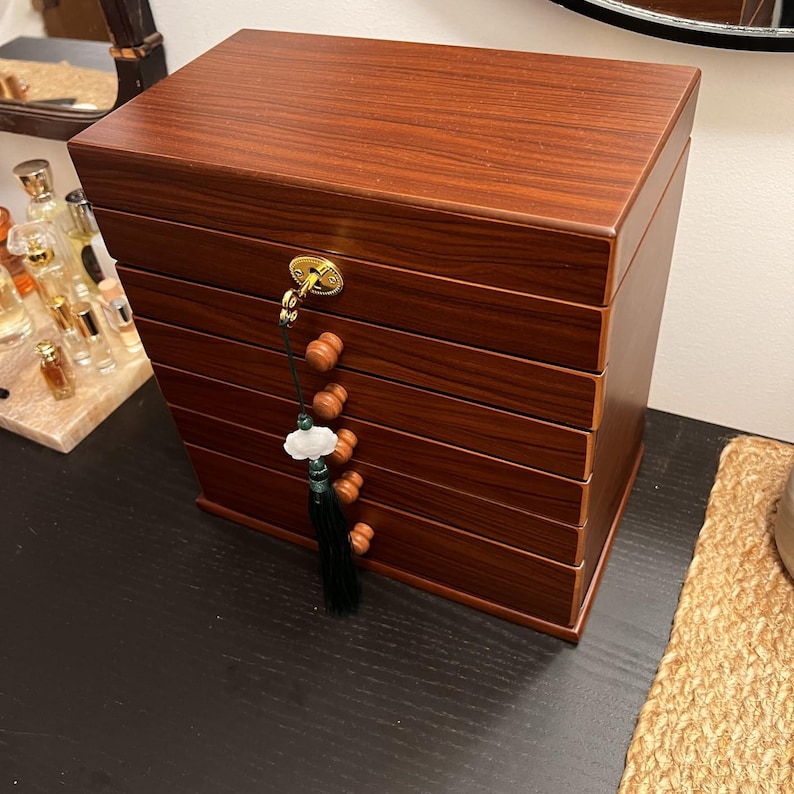 May include: A wooden jewelry box with a rich, reddish-brown finish. The box has multiple drawers and a decorative tassel with wooden beads and a jade-like ornament. A gold-colored clasp secures the lid.