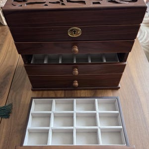 May include: A dark brown wooden jewelry box with multiple drawers and a carved floral design on the top. The drawers have small wooden knobs and are lined with a light-colored fabric. The box is open, revealing the compartments.