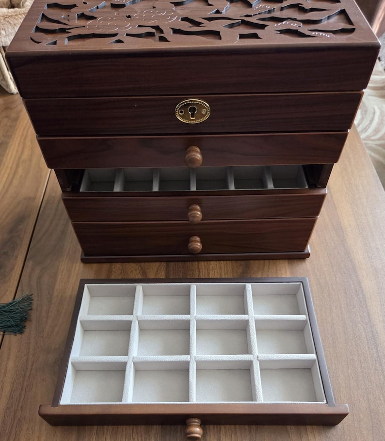 May include: A dark brown wooden jewelry box with multiple drawers and a carved floral design on the top. The drawers have small wooden knobs and are lined with a light-colored fabric. The box is open, revealing the compartments.