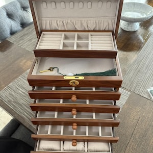 May include: A wooden jewelry box with multiple drawers and compartments. The box is made of dark wood and has a light-colored interior. The top section has ring slots, and the drawers offer various storage options. A gold-colored key and green tassel are visible.