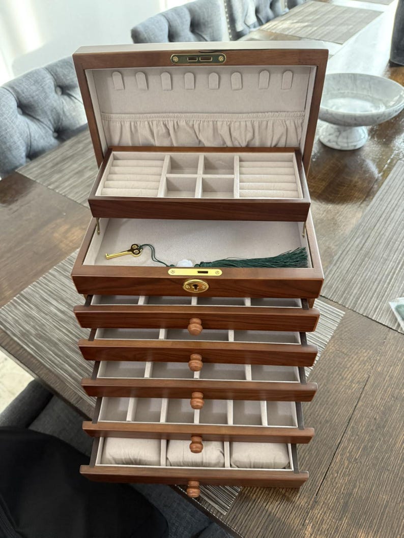 May include: A wooden jewelry box with multiple drawers and compartments. The box is made of dark wood and has a light-colored interior. The top section has ring slots, and the drawers offer various storage options. A gold-colored key and green tassel are visible.