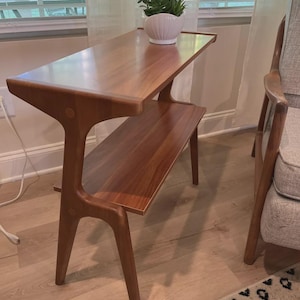May include: A rectangular wooden side table with a smooth, polished finish and a rich brown grain. The table features a lower shelf and angled legs that extend upward to support the top surface. A white, scalloped planter holds a green plant.
