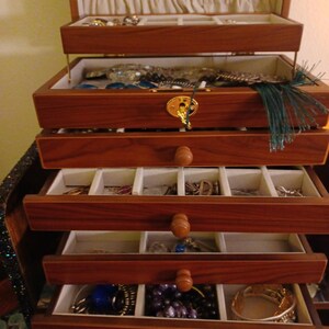 May include: A wooden jewelry box with multiple drawers and compartments. The box is made of dark wood and has a gold-colored clasp. The drawers are lined with a light-colored fabric and contain various pieces of jewelry, including necklaces, bracelets, and earrings.