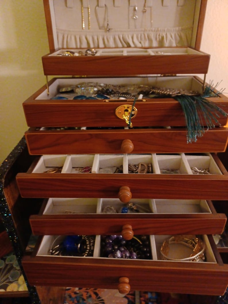 May include: A wooden jewelry box with multiple drawers and compartments. The box is made of dark wood and has a gold-colored clasp. The drawers are lined with a light-colored fabric and contain various pieces of jewelry, including necklaces, bracelets, and earrings.