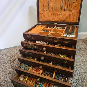 May include: A wooden jewelry box with multiple drawers and compartments, filled with necklaces, bracelets, and earrings. The box is dark brown with floral carvings and a keyhole. The interior is lined with orange fabric.