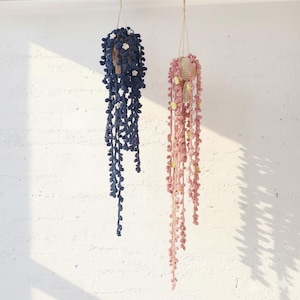 May include: Two hanging crochet plant decorations. One is navy blue with white flower accents, the other is pink with yellow accents. Both have long, trailing strands, resembling string of pearls plants.