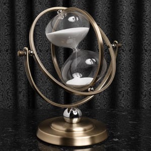 May include: An hourglass with a brass-coloured metal frame and base. The glass bulbs contain white sand. The hourglass is supported by a spherical base and a decorative, swirling frame. The background is a dark, patterned fabric.