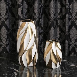 May include: Two decorative vases with a white and gold leaf design. The larger vase is tall and slender, while the smaller one is shorter and wider. Both have a glossy finish and sit on a dark surface.