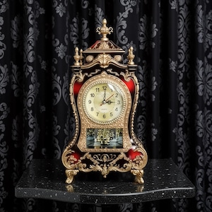 Peut inclure: Une horloge antique décorative avec un cadre doré et un accent rouge. Le cadran de l'horloge est doré avec des chiffres et des aiguilles noirs. L'horloge est ornée de sculptures détaillées et repose sur une surface noire. Mesure environ 30 cm de haut.