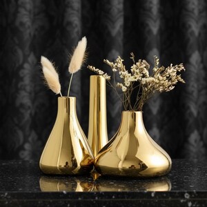 Può includere: Tre vasi color oro di varie forme, con finitura metallica riflettente. Due vasi contengono fiori ed erbe secche. I vasi sono disposti su una superficie scura e strutturata, con una tenda nera a fantasia sullo sfondo.