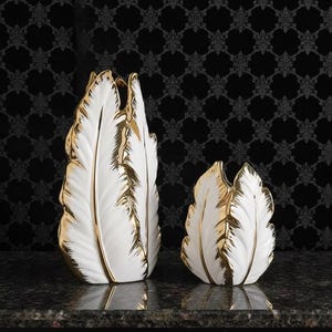 May include: Two white ceramic vases with gold edging, shaped like stylized feathers. The vases are of different sizes and sit on a dark, patterned surface, creating a decorative display.
