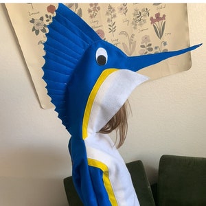 May include: A child wearing a blue and white hooded costume shaped like a marlin fish. The costume has yellow accents and a long, pointed snout.