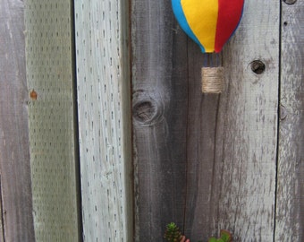 May include: A red, yellow, and blue felt hot air balloon hanging from a wooden fence. Two small potted succulents are in front of the balloon.