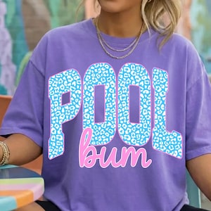 May include: A purple short-sleeve t-shirt featuring the text POOL bum printed on the front. The word POOL is written in large block letters with a light blue and white leopard print pattern, while the word bum is in a pink cursive script.
