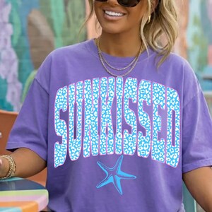以下が含まれることがあります： 紫色のTシャツ。大きな青いヒョウ柄の文字で「SUNKISSED」と書かれています。テキストの下には青いヒトデのグラフィックがあります。画像内の人物はサングラスと金のジュエリーを着用しています。