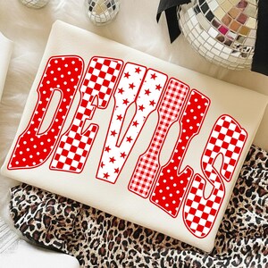 May include: A cream-colored sweatshirt featuring the word DEVILS in large, bold letters. Each letter has a distinct red and white pattern, including polka dots, checkerboard, stars, and gingham prints, with a red outline.