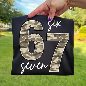 May include: Black t-shirt with the numbers "67" in a camouflage pattern. The word "six" is written above the number 6, and the word "seven" is written below the number 7, both in white script.