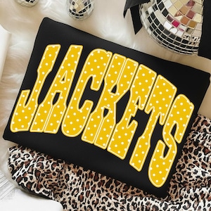 May include: A black sweatshirt featuring the word JACKETS in large, bold, yellow letters. The yellow letters are decorated with a white polka dot pattern and outlined in a thin white border. The text is oriented horizontally across the center.