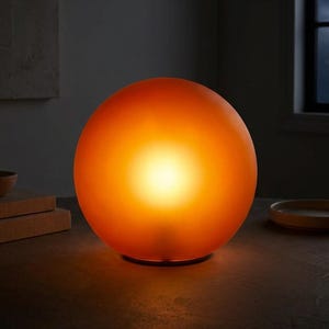 May include: A spherical table lamp with a frosted orange glass shade that emits a warm, glowing light from within. The lamp features a smooth, rounded surface and sits on a circular base, casting a soft illumination across the immediate area.