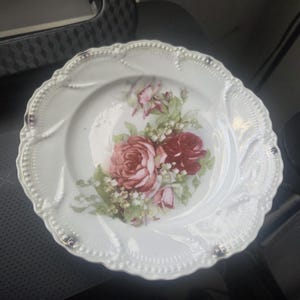 Pode incluir: Prato de porcelana branca com borda ondulada e detalhes em relevo. O prato apresenta um desenho floral central de rosas vermelhas e rosas com folhas verdes. Uma peça decorativa para servir ou expor.