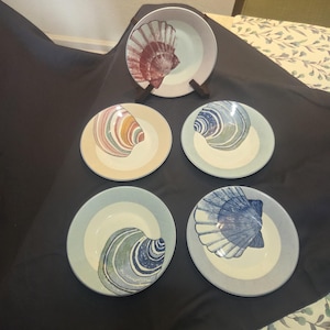 Puede incluir: Un conjunto de cinco platos de cerámica con diseños de conchas marinas. Los platos presentan una variedad de colores, incluyendo rojo, azul, verde y amarillo. Los platos están dispuestos sobre una superficie oscura.