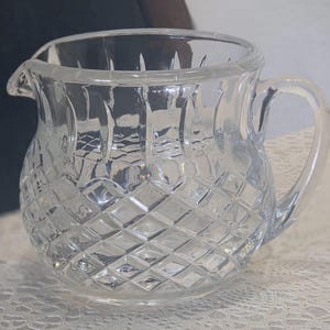 Puede incluir: Una jarra de cristal transparente con un diseño de diamantes. La jarra tiene un pico para verter fácilmente y un asa curvada. Probablemente se utiliza para servir crema o leche.