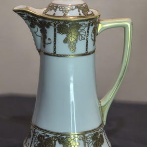Puede incluir: Jarra de cerámica blanca con tapa, con diseños dorados de uvas y hojas. La jarra tiene un asa y un pico curvos, con ribetes dorados. El diseño incluye detalles dorados en la tapa y la base, creando una estética vintage elegante.