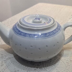 May include: White ceramic teapot with blue floral and geometric patterns. The teapot has a lid with a small floral design and a curved handle. The body of the teapot has small, evenly spaced holes.