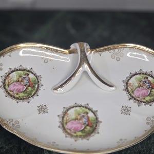 Puede incluir: Plato de porcelana blanco con borde y asa dorados. El plato presenta tres viñetas ovaladas con una pareja con ropa de época. El plato tiene un ribete dorado decorativo y un asa.