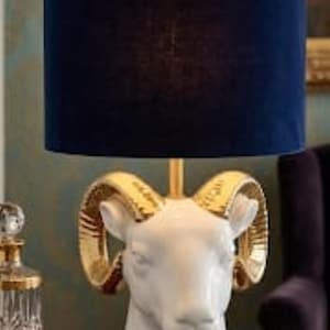 May include: A table lamp featuring a white ceramic ram head base with metallic gold-colored horns and a circular base. The lamp is topped with a cylindrical navy blue velvet lampshade that has a gold-colored trim along the top and bottom edges.